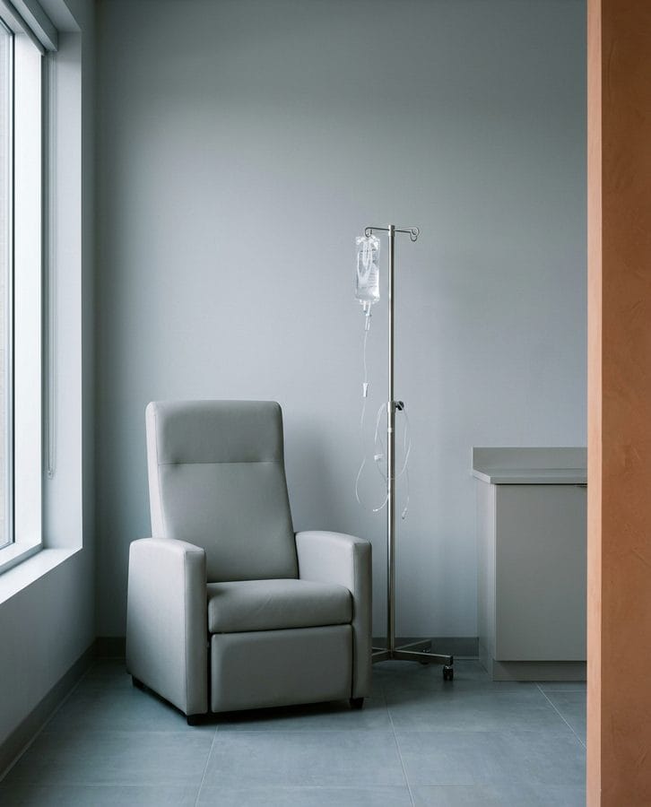 A plain upholstered armchair beside a stainless stand carrying one clear unlabeled fluid bag.