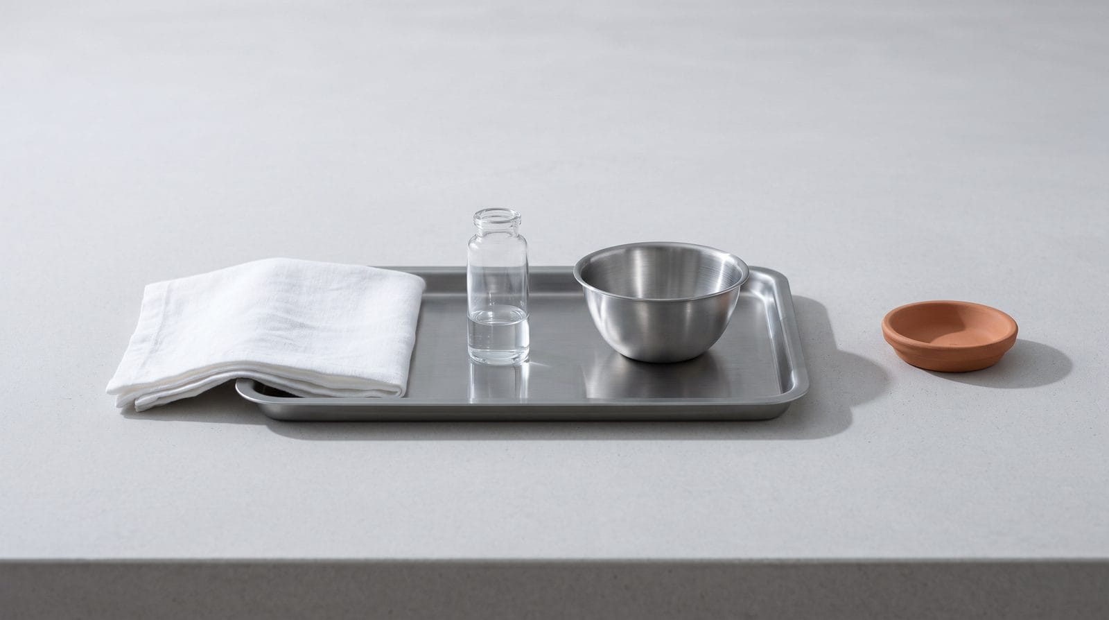 A stainless tray holding one plain unlabeled glass vial and a small stainless mixing bowl.