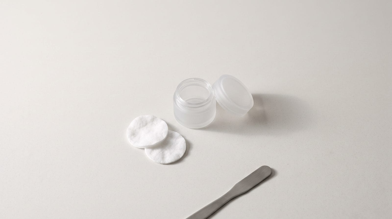 A small frosted glass jar with its lid beside it and two cotton rounds on a pale counter.
